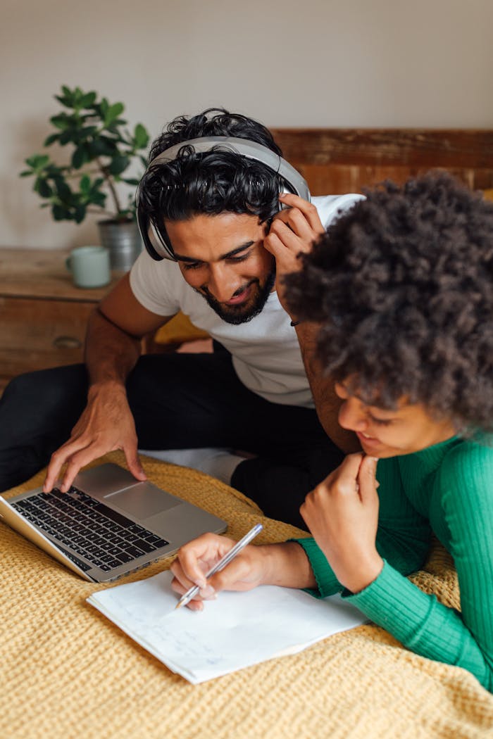 Two young adults working together at home, using a laptop and taking notes for a project.
